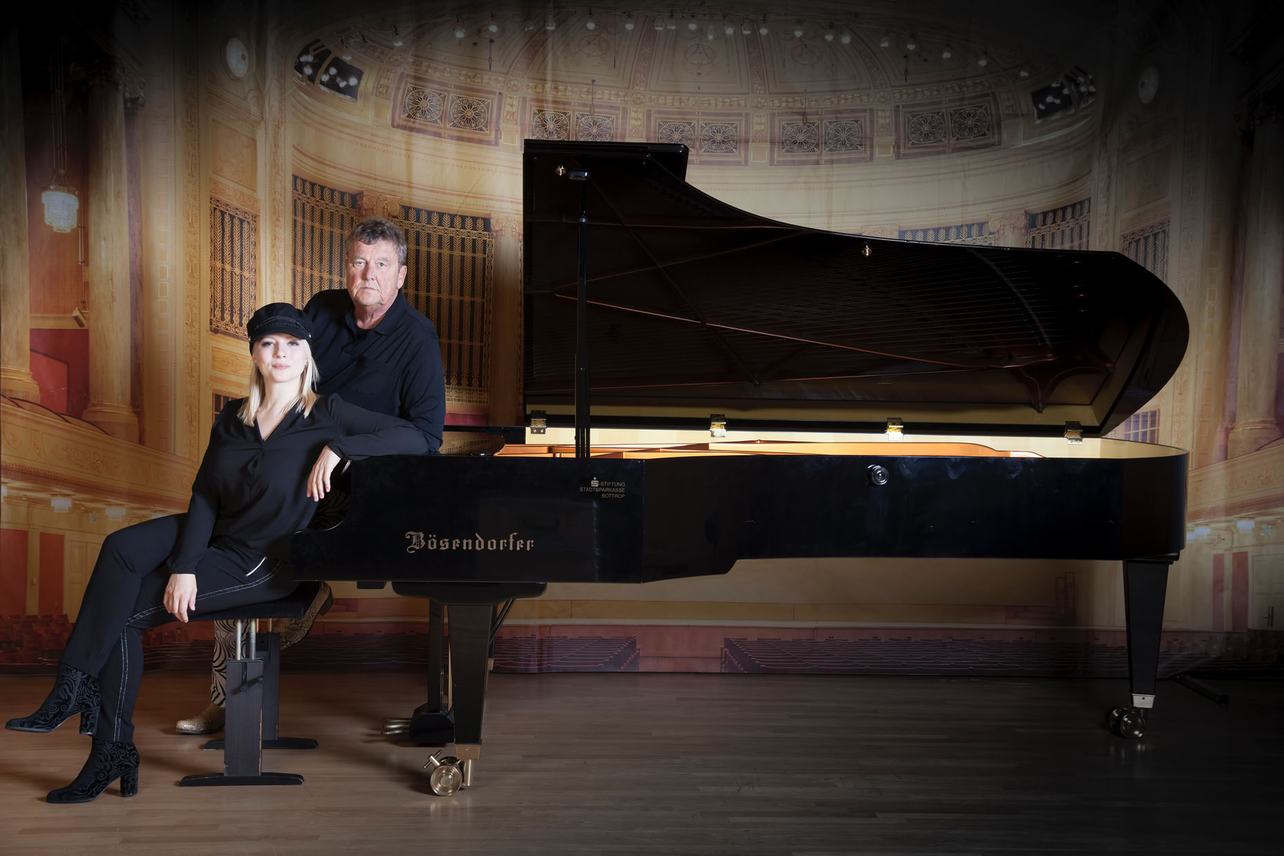 Nadia Singer und Lutz G&ouml;rner in schwarzer Kleidung sitzen auf einem Hocker vor einem gro&szlig;en Fl&uuml;gel in einem Konzertsaal.