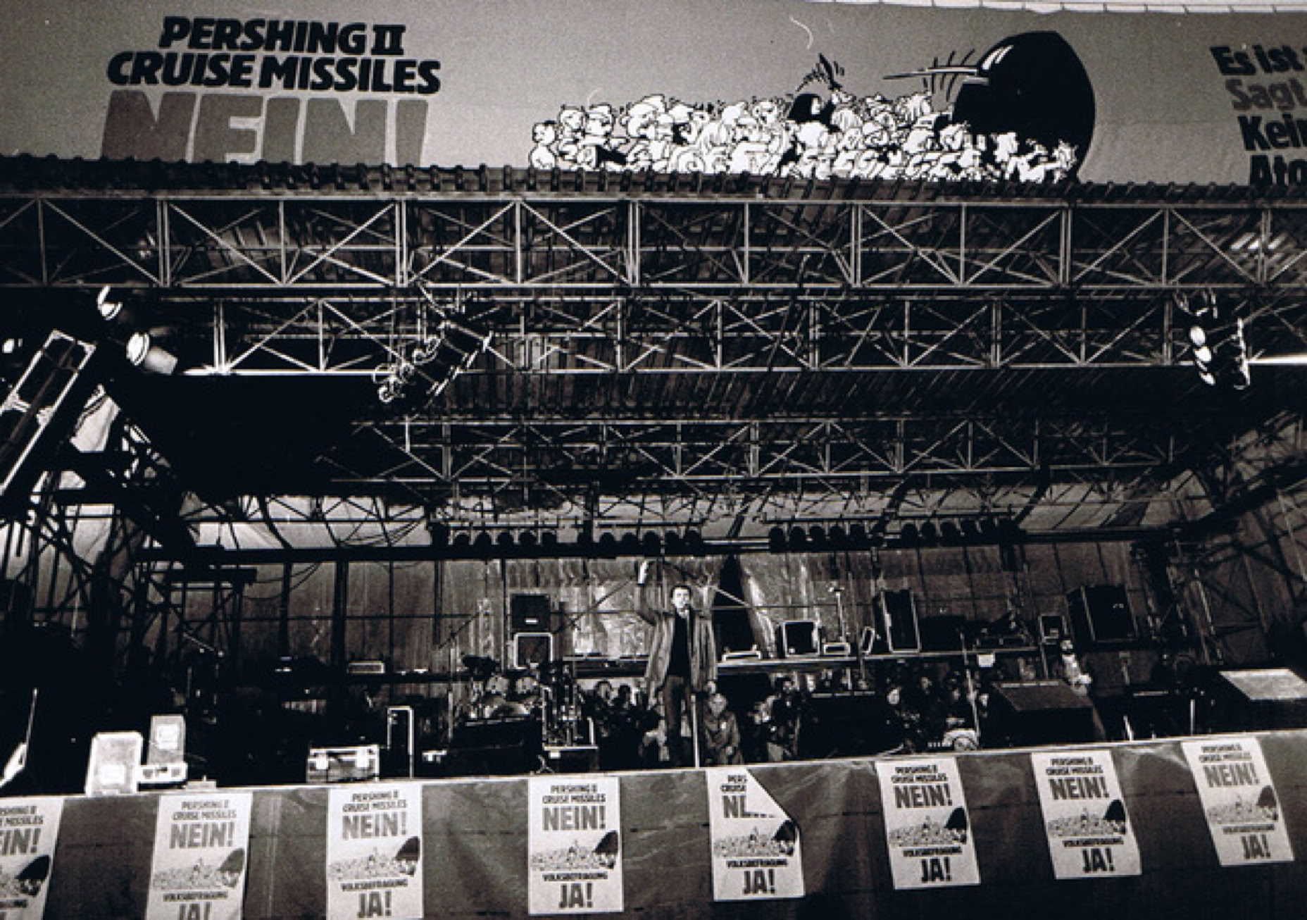 Lutz G&ouml;rner Vita: Eine B&uuml;hne mit Mikrofonen und Instrumenten, im Hintergrund ein gro&szlig;es Plakat mit der Aufschrift "PERSHING II CRUISE MISSILES NEIN!" und mehreren kleineren Plakaten im Vordergrund. Friedensdemo im Bonner Hofgarten.