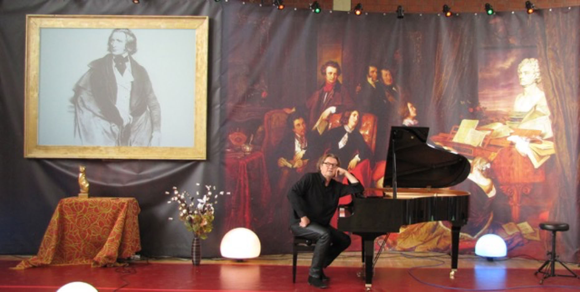 Lutz G&ouml;rner sitzt an einem Fl&uuml;gel auf einer B&uuml;hne mit einem gro&szlig;en Gem&auml;lde im Hintergrund und einem gerahmten Bild von Franz Liszt an der Wand.